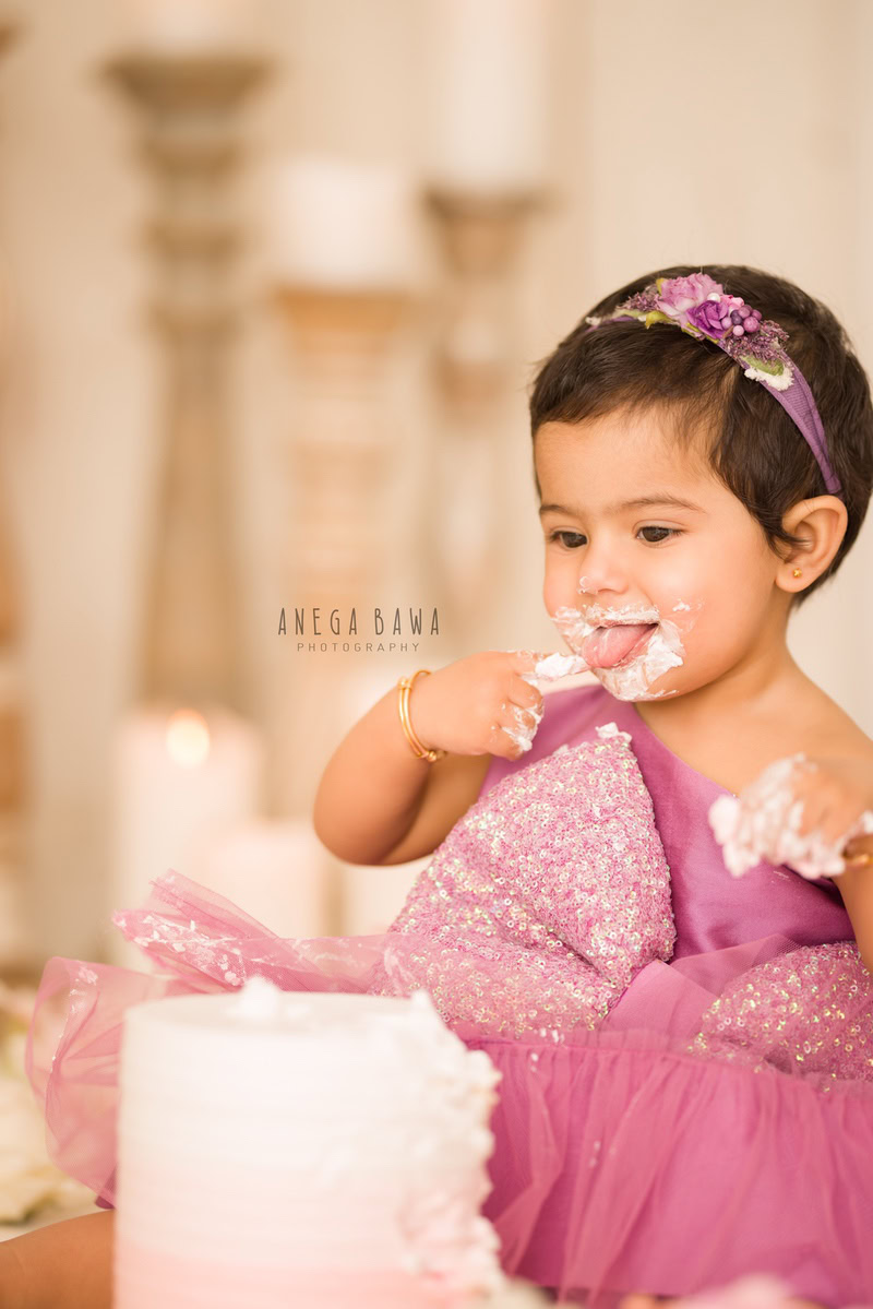 Girl with cute headband, cake smash on face, candle stand, beige backdrop. First birthday photography in Delhi, Gurgaon. Captured by Anega Bawa family photographer Gurgaon (Delhi NCR).