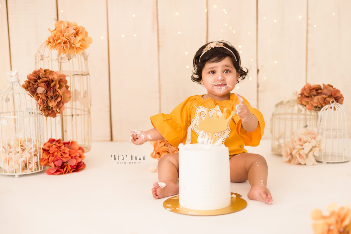 Girl in a yellow dress enjoying a Cake Smash with a beige backdrop featuring white castles during a first birthday photoshoot by well-known Anega Bawa Photography in Delhi, Gurgaon.
