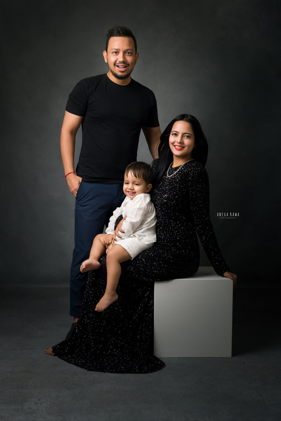 1 year boy sitting on mom’s lap with dad posing beside them, set against a grey backdrop in a black-and-white family photography session by Anega Bawa in Delhi, Gurgaon. The classic tones highlight their family bond.