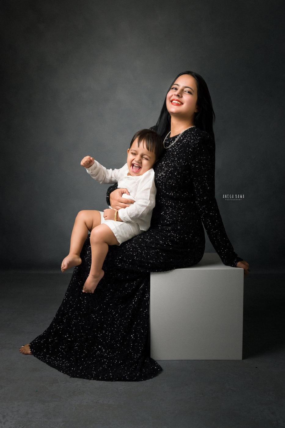 1 year boy sitting on mom’s lap in a cute smiling pose against a grey backdrop, captured during a family photography session by Anega Bawa in Delhi, Gurgaon. Their joyful expressions create a heartwarming moment.