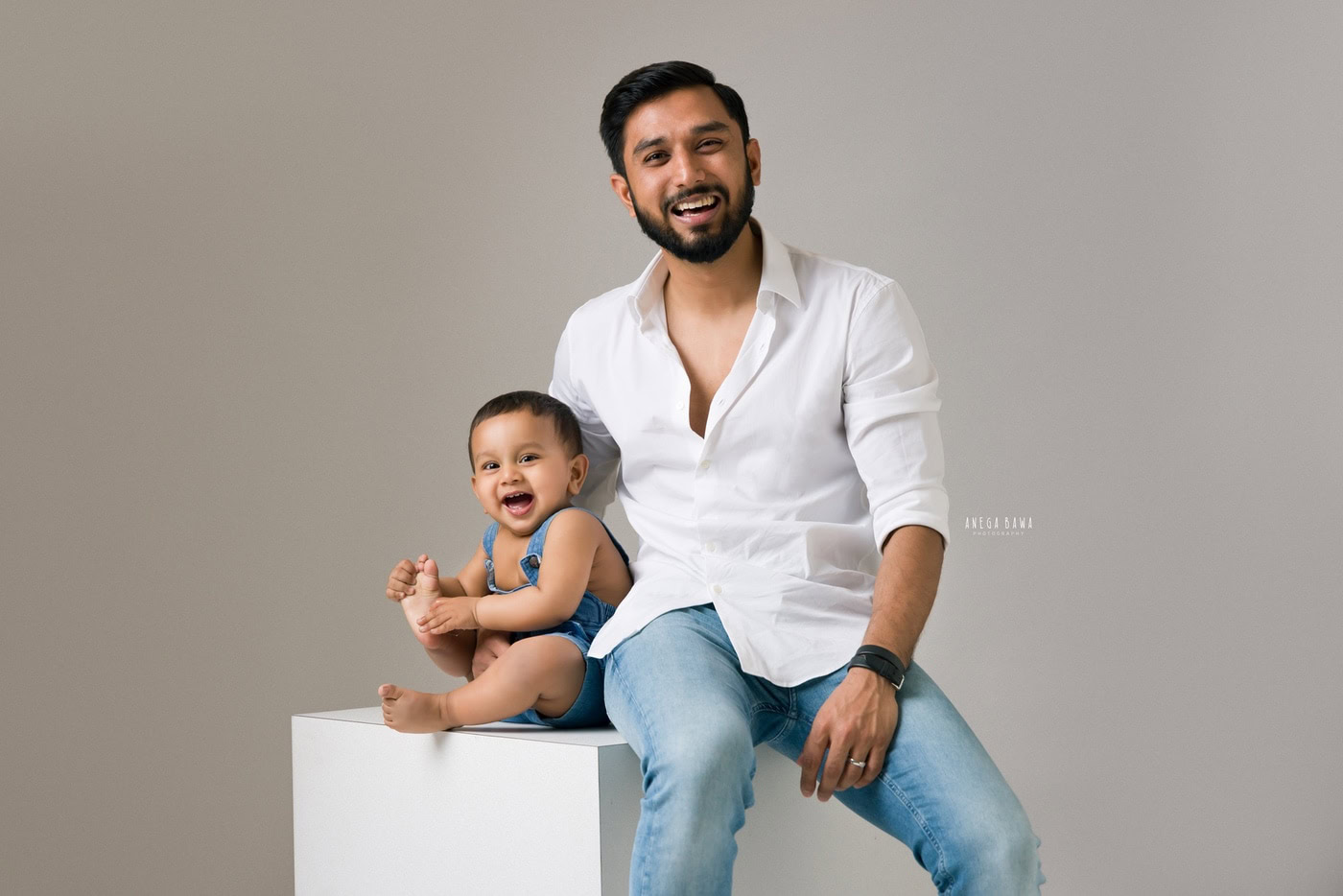 1 year boy wearing a denim dungaree, sitting on a white stool with dad in a smiling pose, captured against a grey backdrop during a family photography session by Anega Bawa in Delhi, Gurgaon. The casual setting showcases their joyful connection.