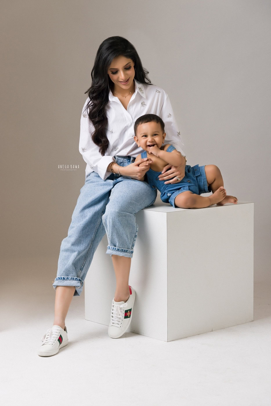 1 year boy wearing a denim dungaree, sitting on a white stool with mom dressed in a white shirt and denims, set against a grey backdrop, captured during a family photography session by Anega Bawa in Delhi, Gurgaon. The coordinated outfits add a harmonious touch to the scene.