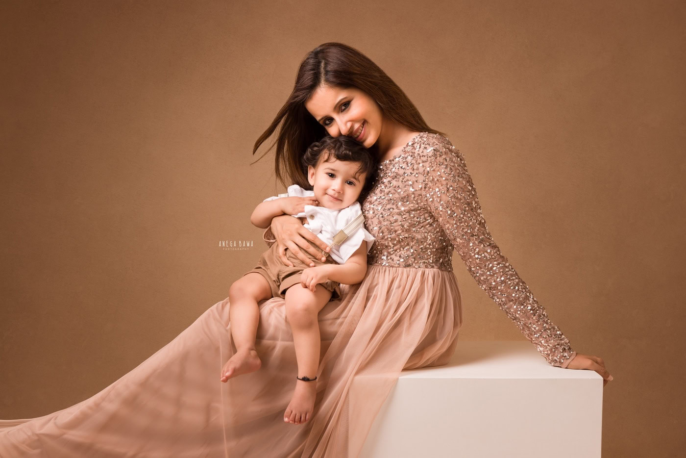 1 year girl posing with mom wearing a peach gown against a brown backdrop, captured during a family photography session by Anega Bawa in Delhi, Gurgaon. The elegant setting highlights their bond.