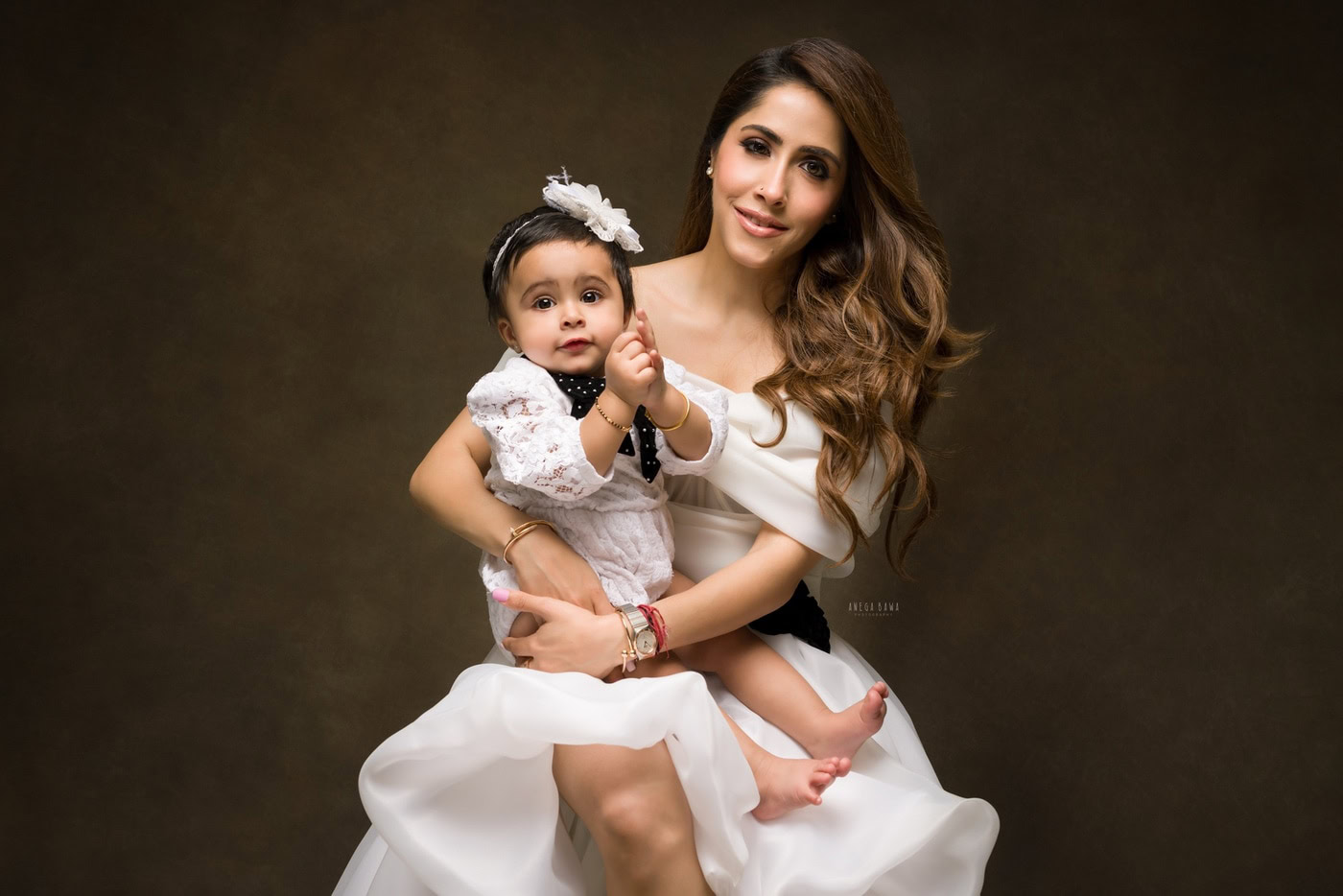 1 year girl sitting on mom’s lap, wearing a cute headband against a brown backdrop, captured during a family photography session by Anega Bawa in Delhi, Gurgaon. The warm tones enhance the tenderness of their connection.