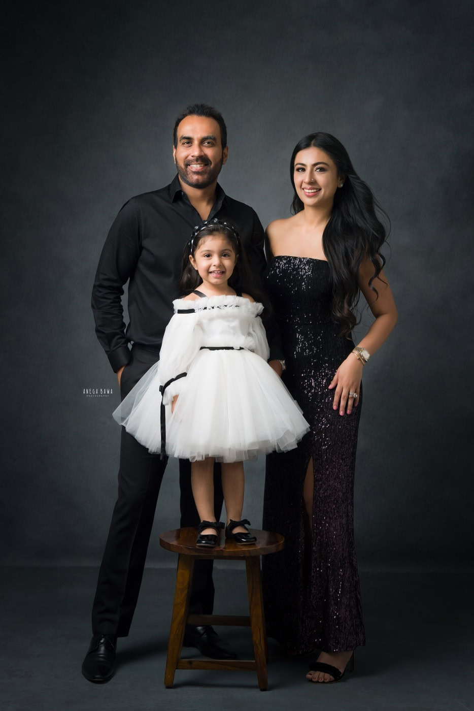 1-year-old girl wearing a white dress, posing with her mom and dad against a grey backdrop, captured during a family photography session by Anega Bawa in Delhi, Gurgaon. Their joyful expressions and loving poses showcase the warmth of their family bond.