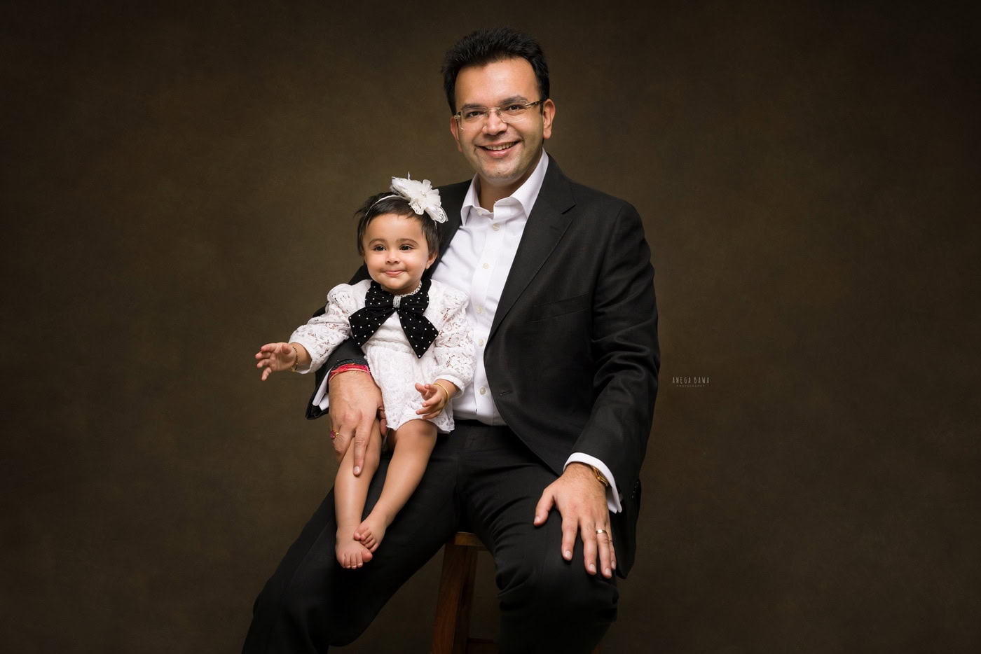 1 year girl wearing a white dress, sitting on dad’s lap against a brown backdrop, captured during a family photography session by Anega Bawa in Delhi, Gurgaon. The contrasting colors highlight their loving bond.
