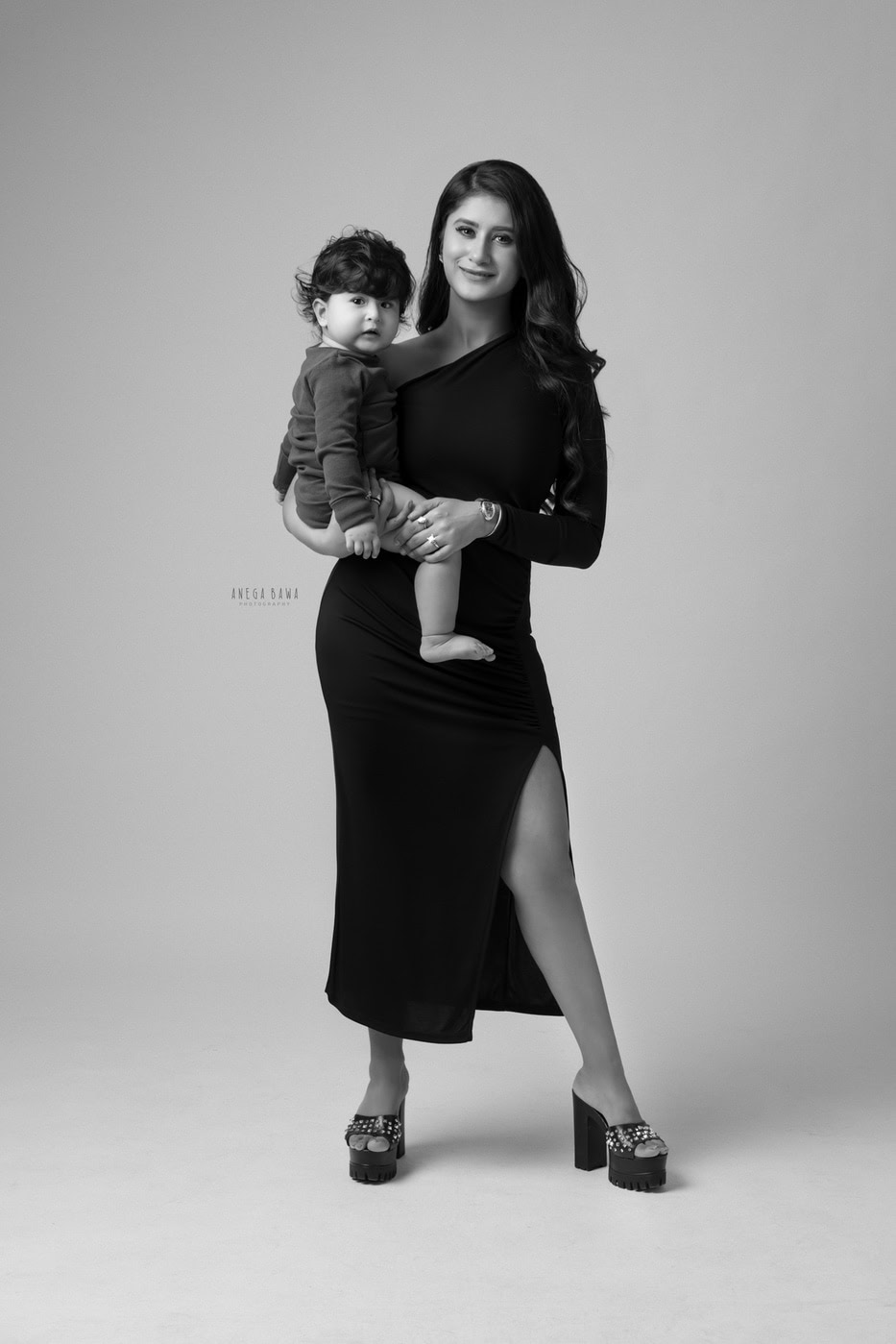 12 month boy in mom's arms against a grey backdrop in a black-and-white family photography session by Anega Bawa in Delhi, Gurgaon. The classic tones highlight their close bond and tender moment.