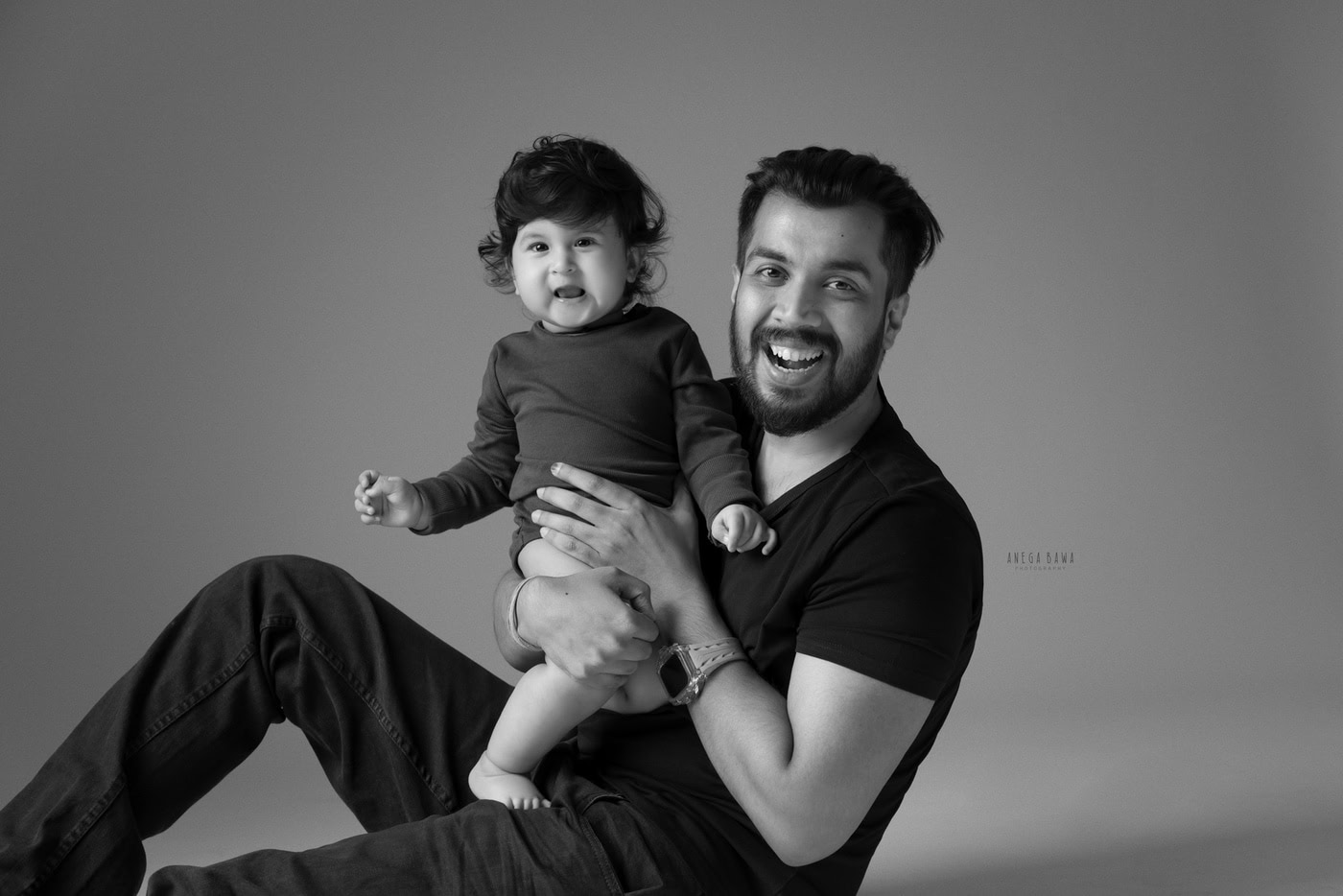 12 month boy posing cutely with dad against a grey backdrop, captured during a family photography session by Anega Bawa in Delhi, Gurgaon. Their joyful expressions create a heartwarming moment.