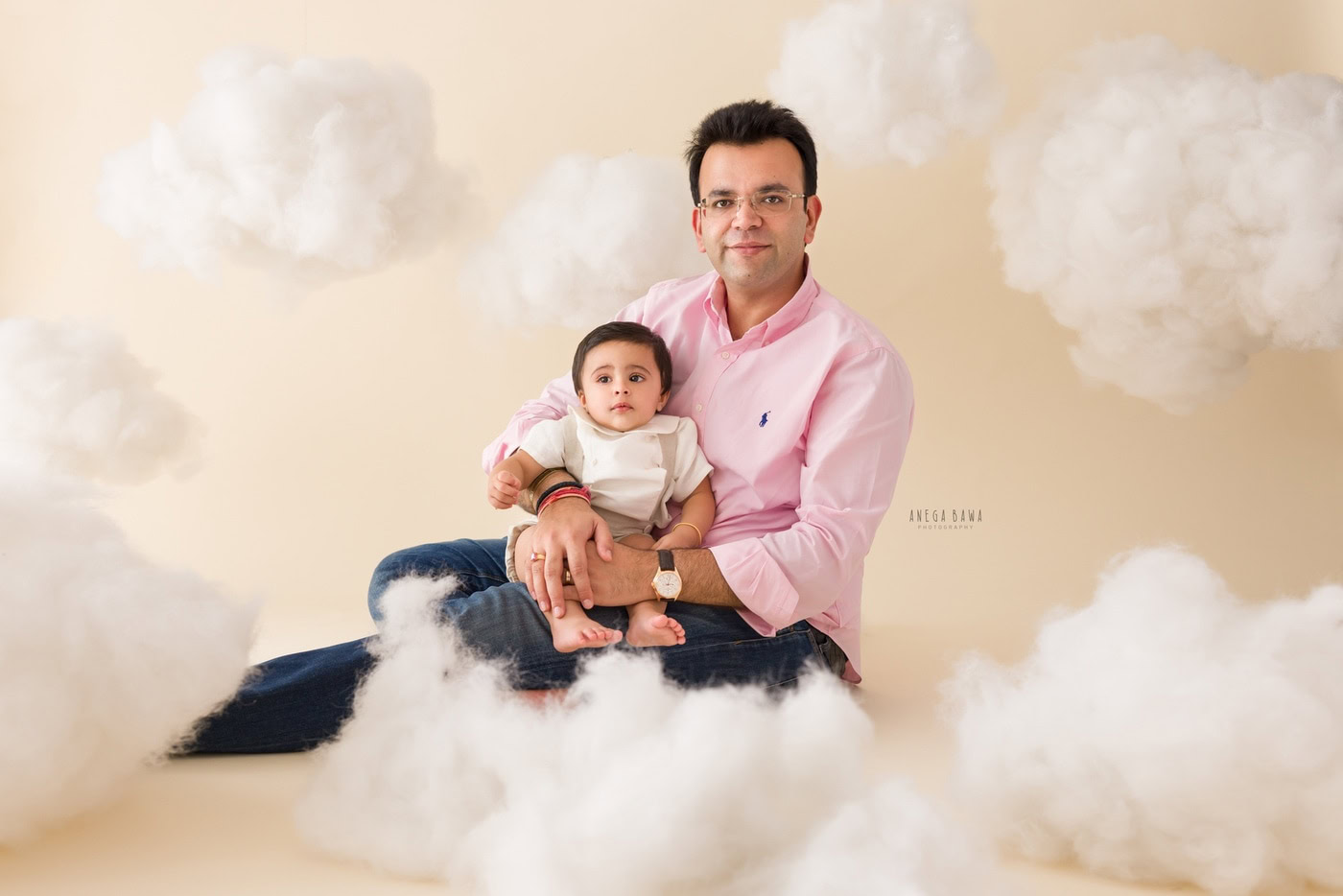 12 month boy sitting on dad's lap in a cute pose against a beige cloudy backdrop, captured during a family photography session by Anega Bawa in Delhi, Gurgaon. Their playful interaction highlights the special bond they share.
