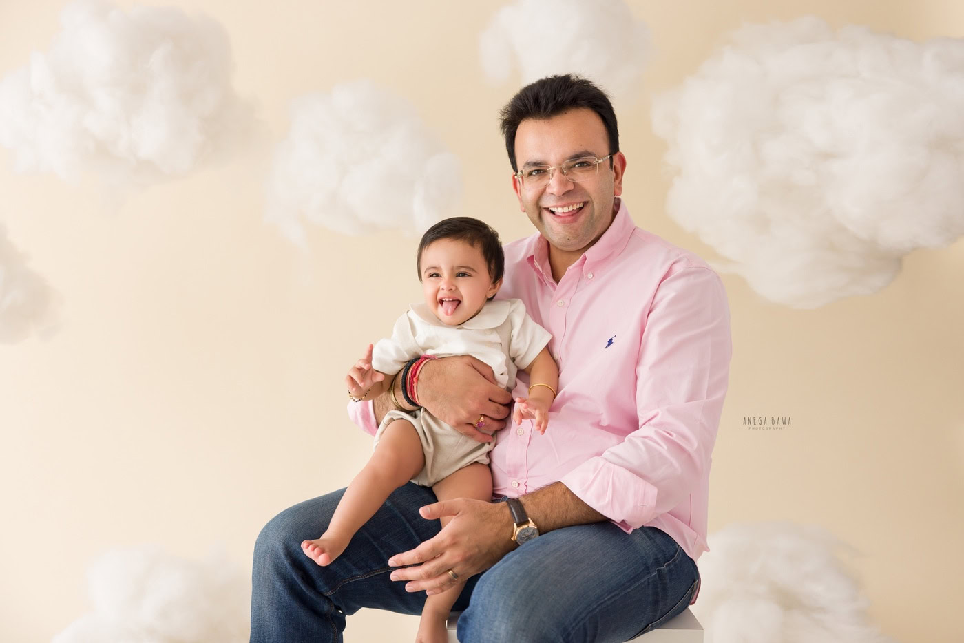 12 month boy sitting on dad's lap against a beige cloudy backdrop, captured during a family photography session by Anega Bawa in Delhi, Gurgaon. This heartwarming moment showcases their close relationship and joyful expressions.