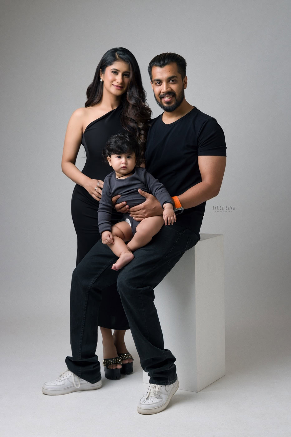 12 month boy sitting on dad's lap, posing with mom against a white backdrop, captured during a family photography session by Anega Bawa in Delhi, Gurgaon. Their happy expressions highlight the warmth of their family bond.