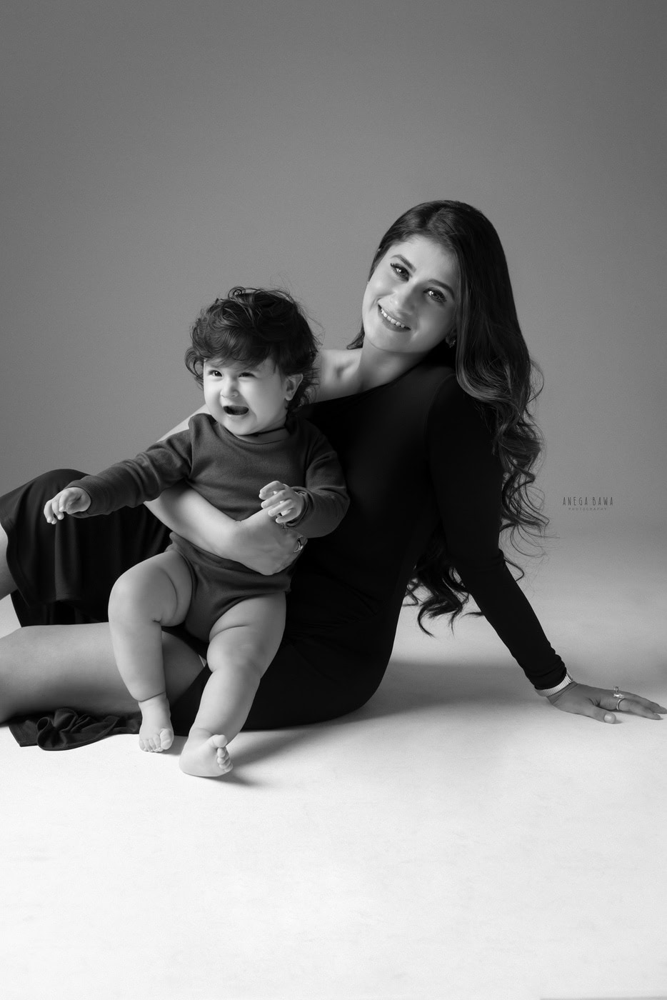 12 month son posing cutely with mom, sitting on the floor against a grey backdrop in a black-and-white family photography session by Anega Bawa in Delhi, Gurgaon. Their joyful expressions highlight the warmth of their bond.