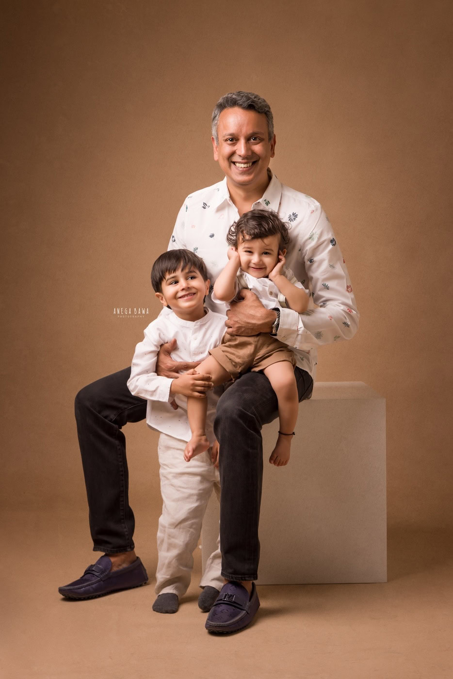 12 month son sitting on dad's lap, posing with his elder brother against a light brown backdrop, captured during a family photography session by Anega Bawa in Delhi, Gurgaon. Their happy expressions showcase the strong sibling bond and family connection.