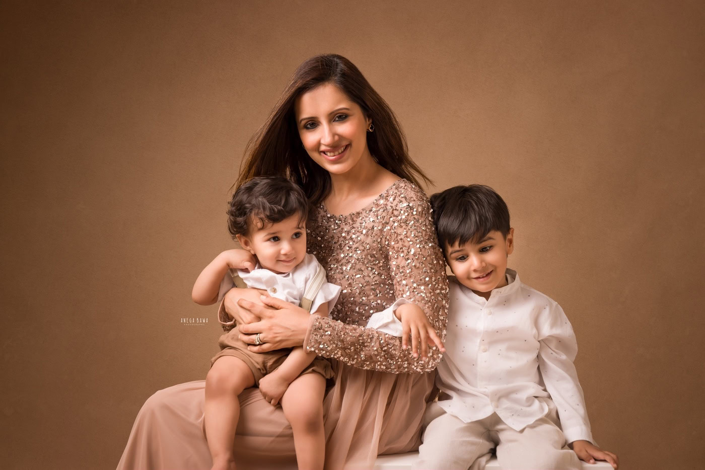12 month son sitting on mom's lap, posing with his elder brother against a brown backdrop, captured during a family photography session by Anega Bawa in Delhi, Gurgaon. Their joyful expressions highlight the loving family connection.