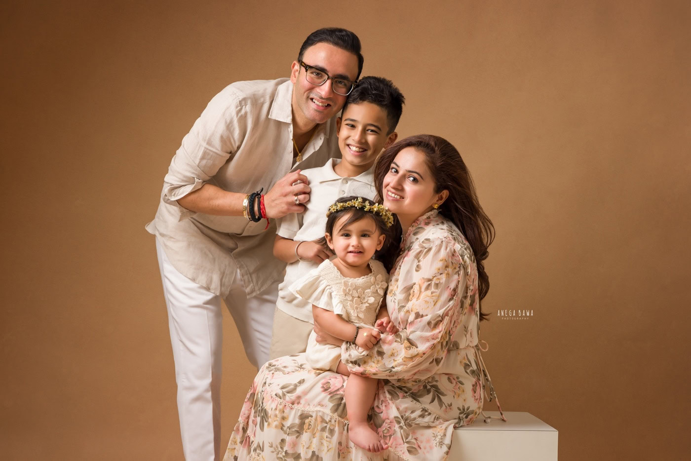 15-month-old daughter sitting on her mom's lap, posing with dad and her elder brother against a light brown backdrop, captured during a family photography session by Anega Bawa in Delhi, Gurgaon. Their warm smiles and loving interaction highlight the family's joyful bond.