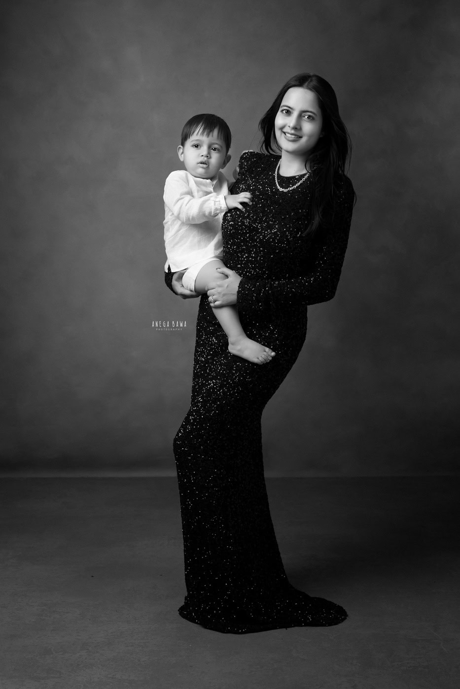 15 month son posing with mom in a black gown against a grey backdrop in a black-and-white family photography session by Anega Bawa in Delhi, Gurgaon. The classic tones enhance their elegant connection and joyful expressions.