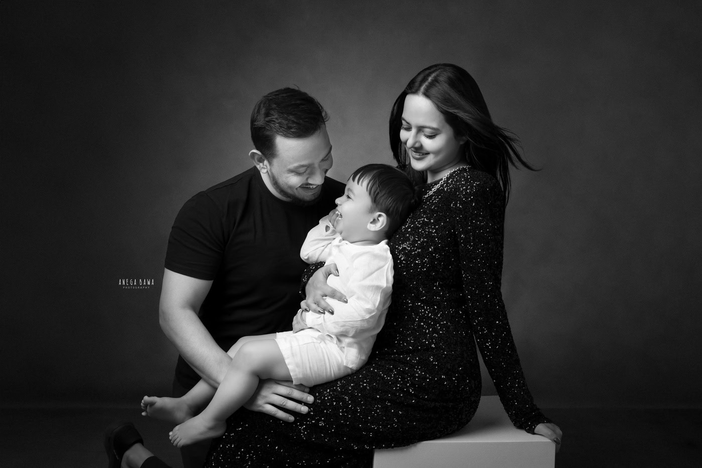 2 year boy posing cutely with mom and dad against a grey backdrop in a black-and-white family photography session by Anega Bawa in Delhi, Gurgaon. The timeless style enhances their family bond and joyful expressions.