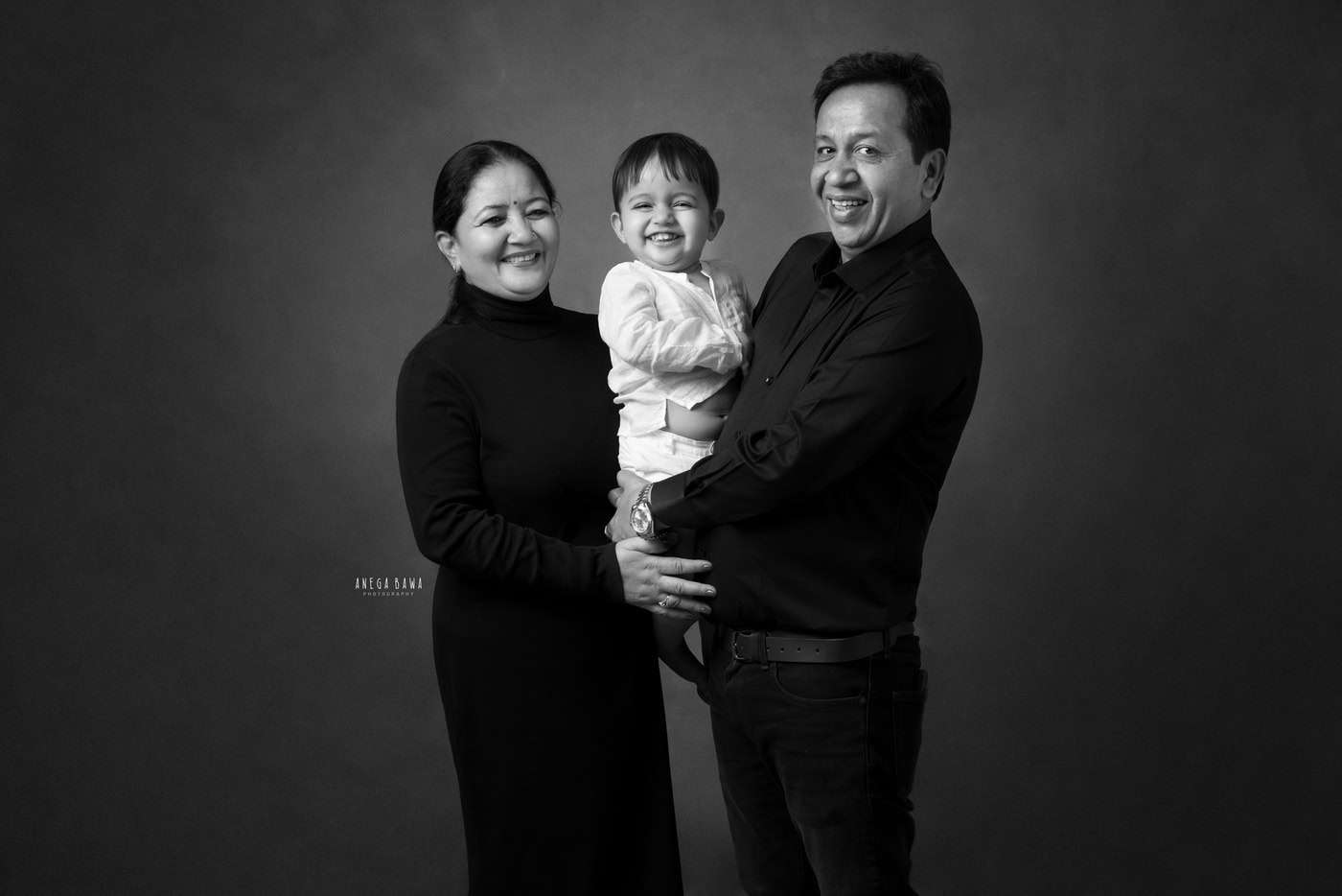 2 year boy posing with his grandparents against a grey backdrop, captured during a family photography session by Anega Bawa in Delhi, Gurgaon. The warm interaction highlights the loving family connection.