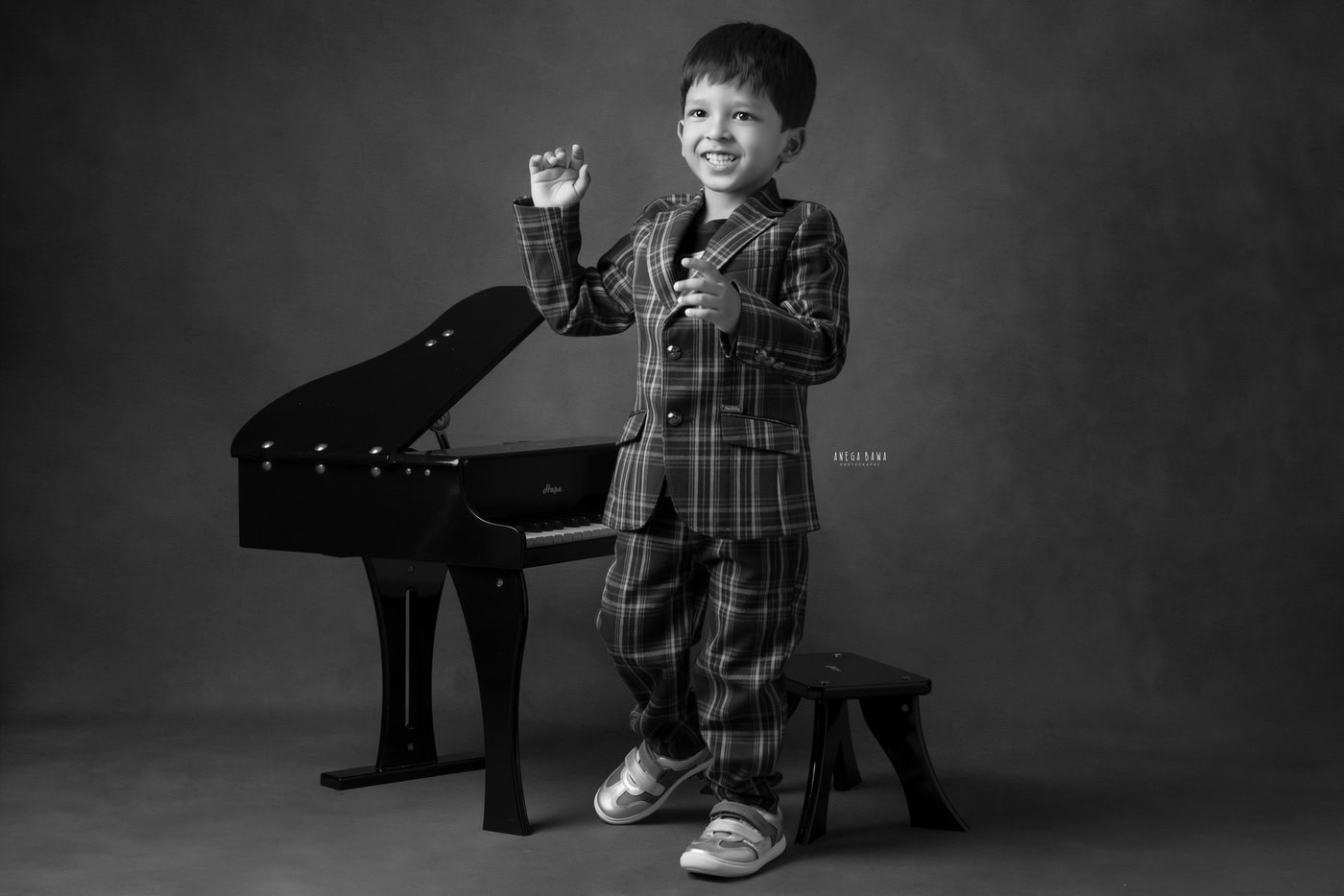 2-year-old boy posing with a piano in a cute smiling pose against a black-and-white backdrop, captured during a photography session by Anega Bawa in Delhi, Gurgaon. His playful expression beautifully showcases his charm and personality.