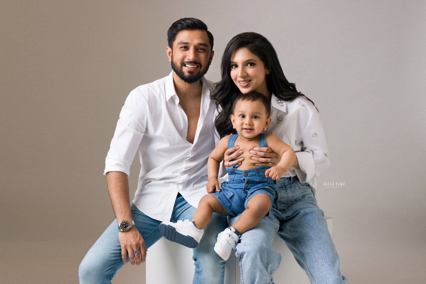2 year boy wearing a denim dungaree, posing cutely with mom and dad against a grey backdrop, captured during a family photography session by Anega Bawa in Delhi, Gurgaon. Their cheerful expressions create a heartwarming family moment.