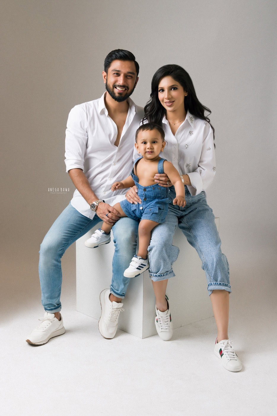 2 year boy wearing a denim dungaree, sitting on mom's lap and posing with dad in a white shirt against a grey backdrop, captured during a family photography session by Anega Bawa in Delhi, Gurgaon. The playful scene highlights their joyful family dynamic.