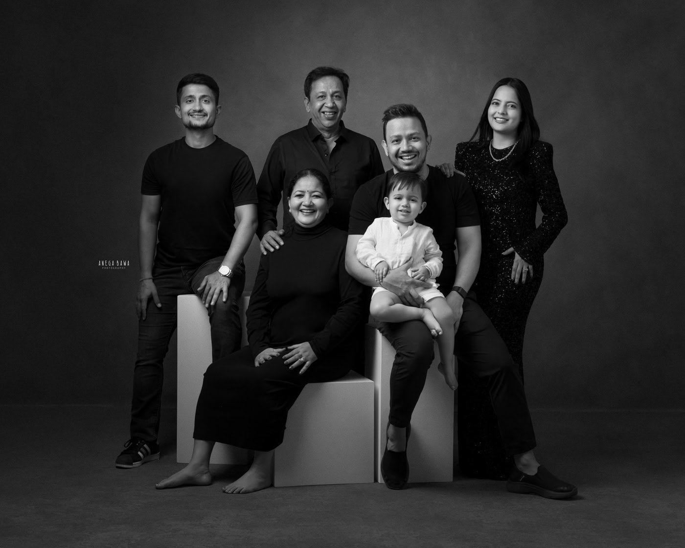 2 year boy wearing a white shirt, sitting on dad's lap and posing with mom, grandparents, and uncle, all in black against a black-and-white backdrop, captured during a family photography session by Anega Bawa in Delhi, Gurgaon. The striking contrast enhances their loving family connection.