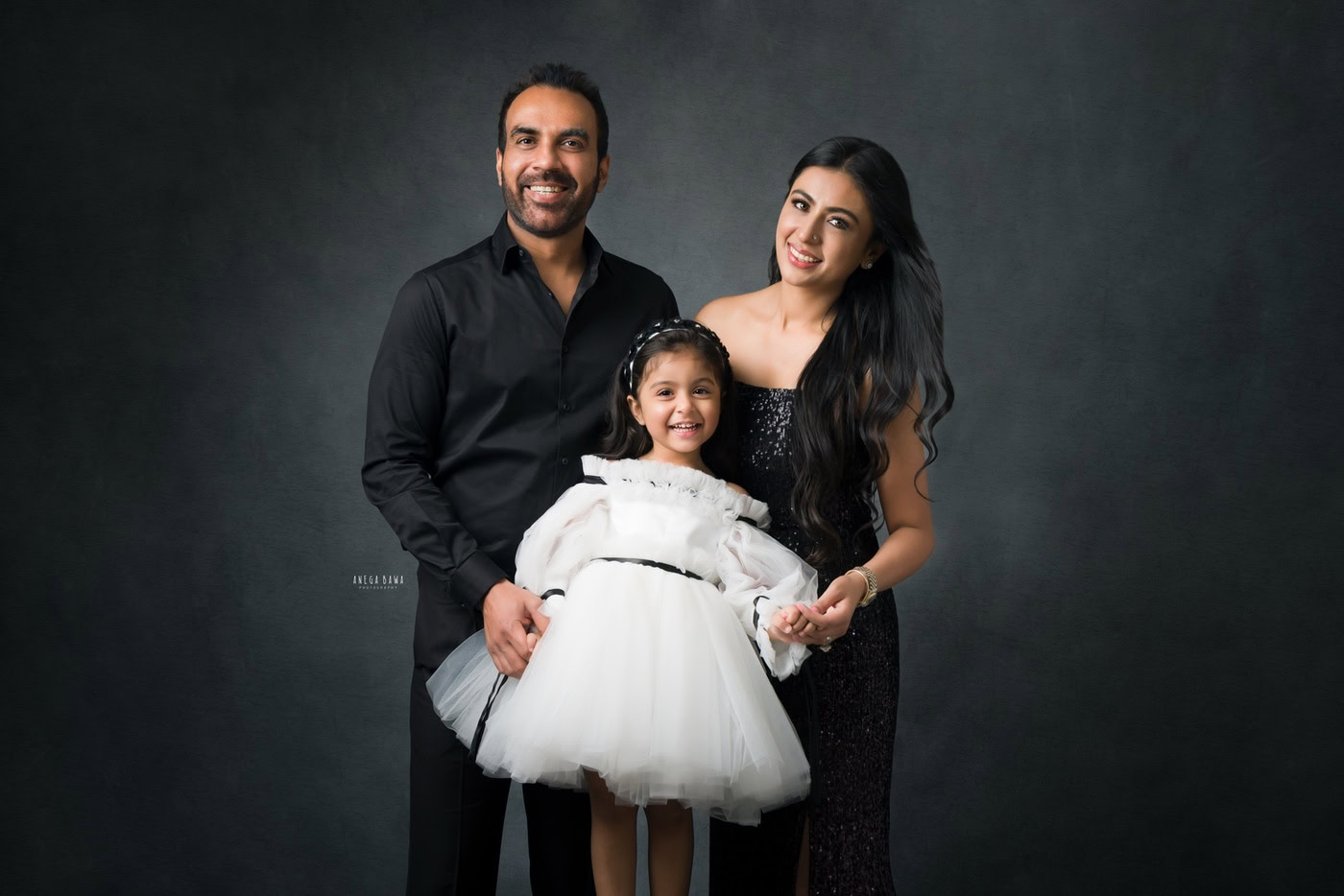 2-year-old daughter posing cutely with her mom and dad against a grey backdrop, captured during a family photography session by Anega Bawa in Delhi, Gurgaon. Their joyful smiles and affectionate poses highlight the love within their family.
