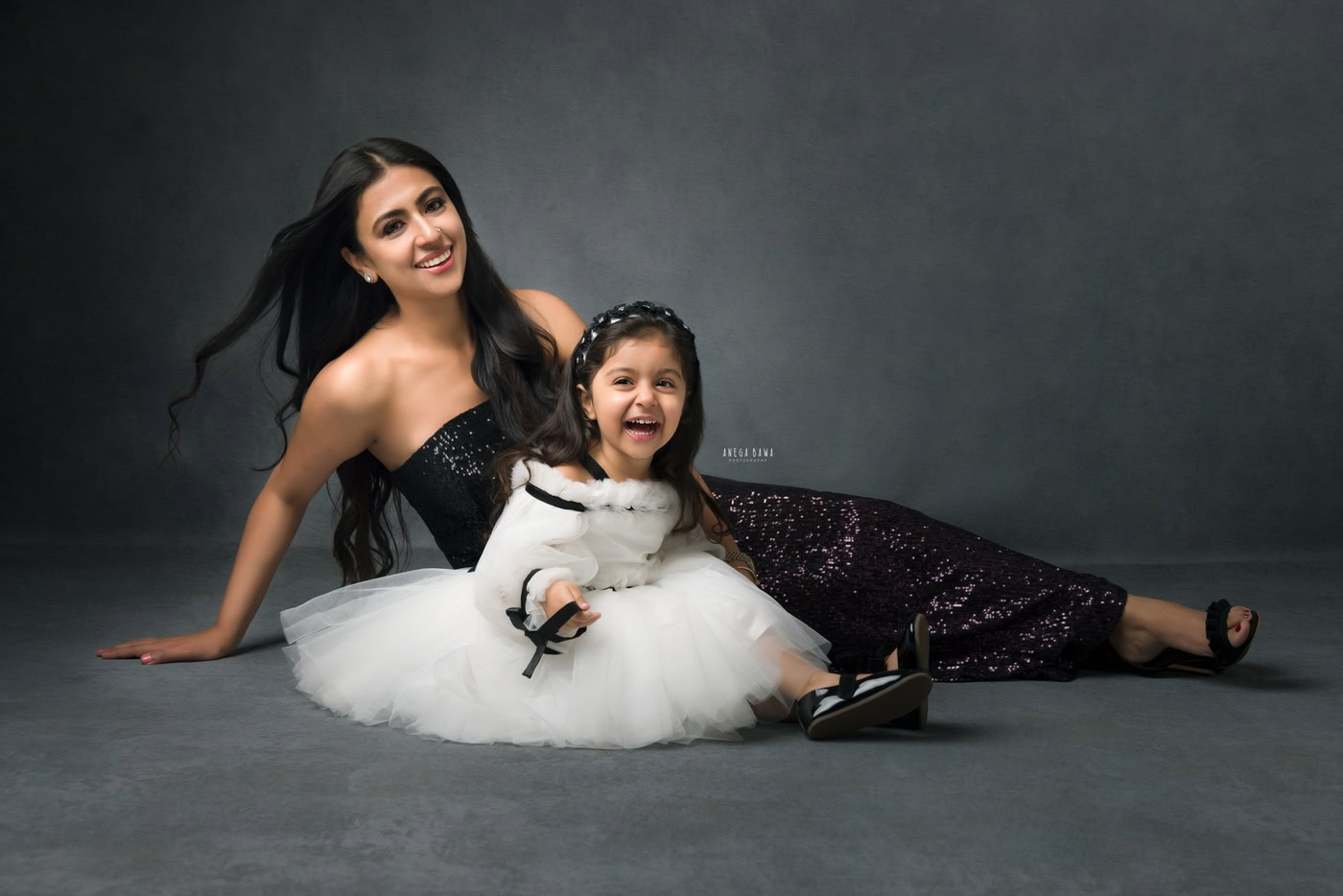 2-year-old girl sitting on the floor in a white dress, posing with her mom against a grey backdrop, captured during a family photography session by Anega Bawa in Delhi, Gurgaon. Their warm smiles and playful poses beautifully showcase their loving bond.