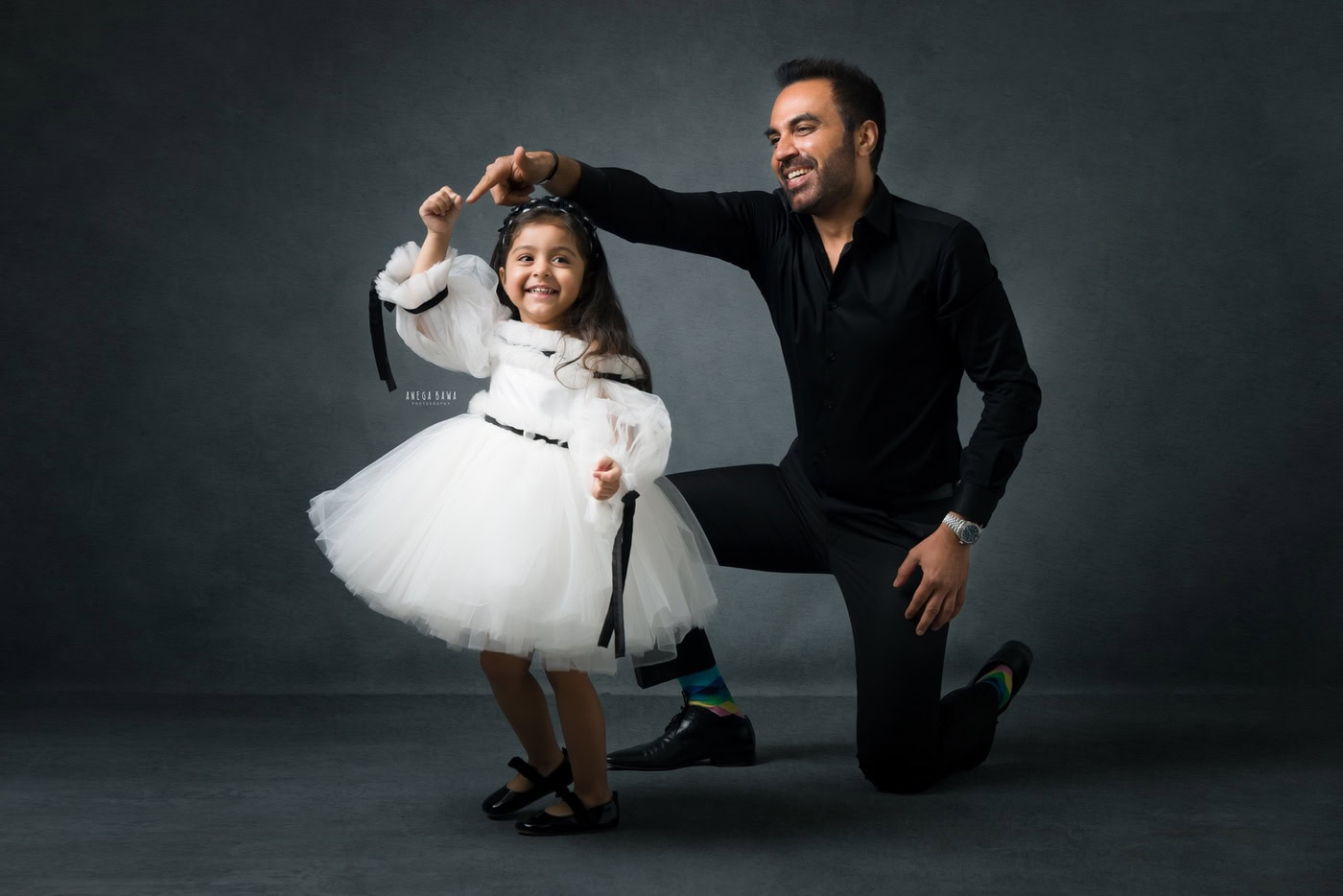 2-year-old girl wearing a white dress, posing with her dad in a dancing pose against a grey backdrop, captured during a family photography session by Anega Bawa in Delhi, Gurgaon. Their joyful expressions and playful interaction highlight the fun and love in their relationship.