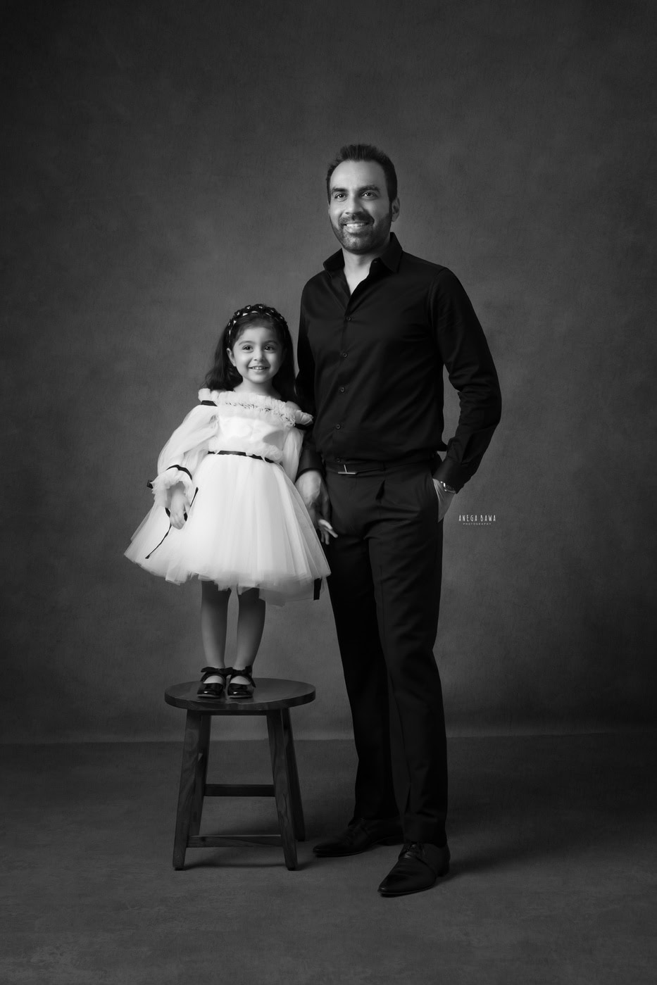 2-year-old girl wearing a white dress, standing on a wooden stool and posing with her dad against a black-and-white backdrop, captured during a family photography session by Anega Bawa in Delhi, Gurgaon. Their playful interaction and bright smiles beautifully showcase their bond.