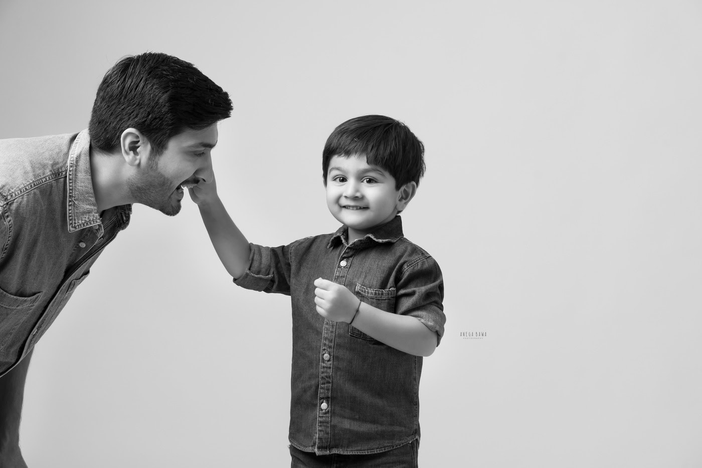 3 year son playing with dad in a cute pose against a grey backdrop, captured during a family photography session by Anega Bawa in Delhi, Gurgaon. Their playful interaction highlights the joy of their bond.
