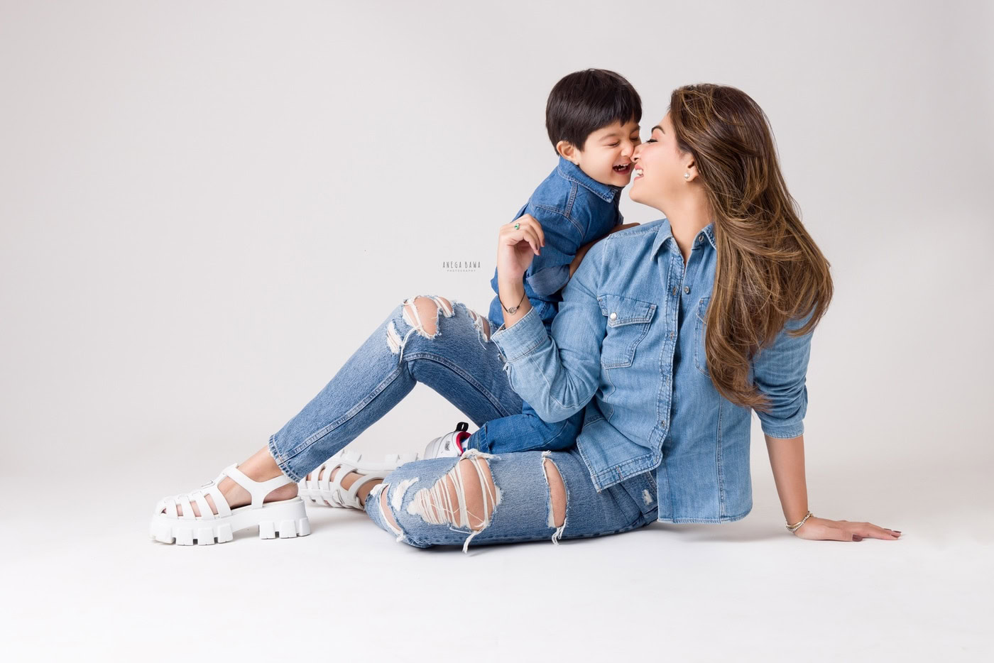 3 year boy in a cute pose with mom sitting on the floor against a white backdrop, captured during a family photography session by Anega Bawa in Delhi, Gurgaon. Their playful expressions create a warm and joyful moment.