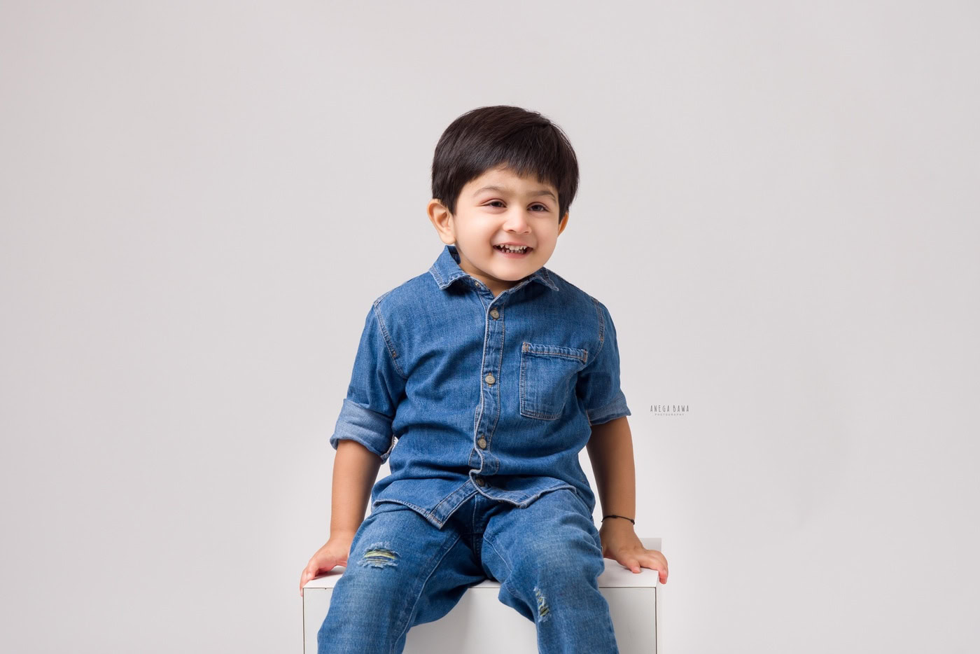 3 year boy posing cutely in an all-denim look against a white backdrop, captured during a family photography session by Anega Bawa in Delhi, Gurgaon. The clean background highlights his playful personality and style.