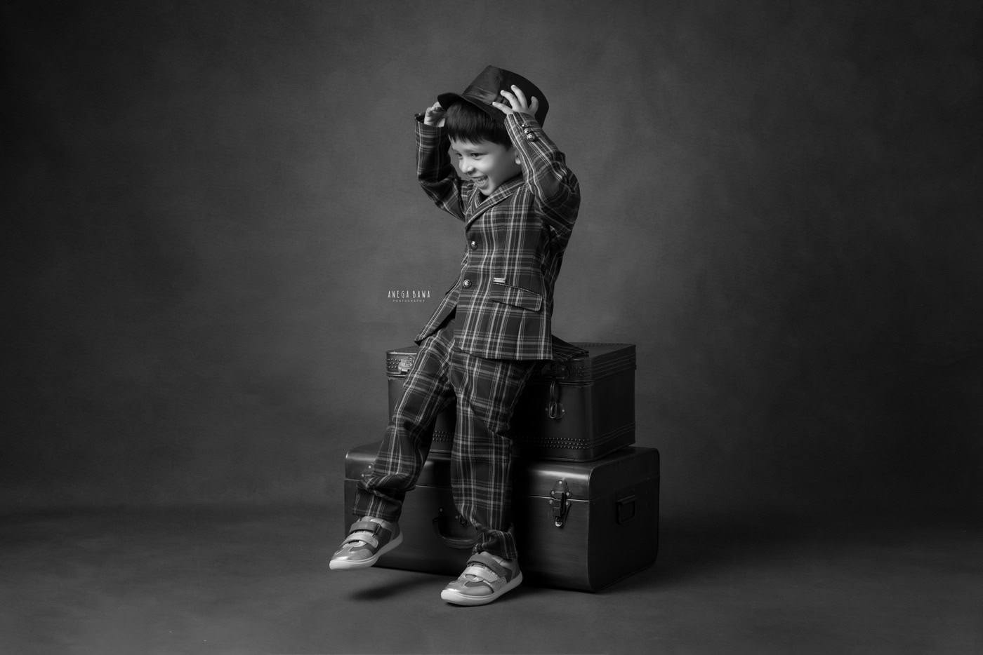 3-year-old boy posing in a check suit, playfully wearing a hat on his head against a black-and-white backdrop, captured during a family photography session by Anega Bawa in Delhi, Gurgaon. His playful demeanor and charming outfit highlight his fun personality.