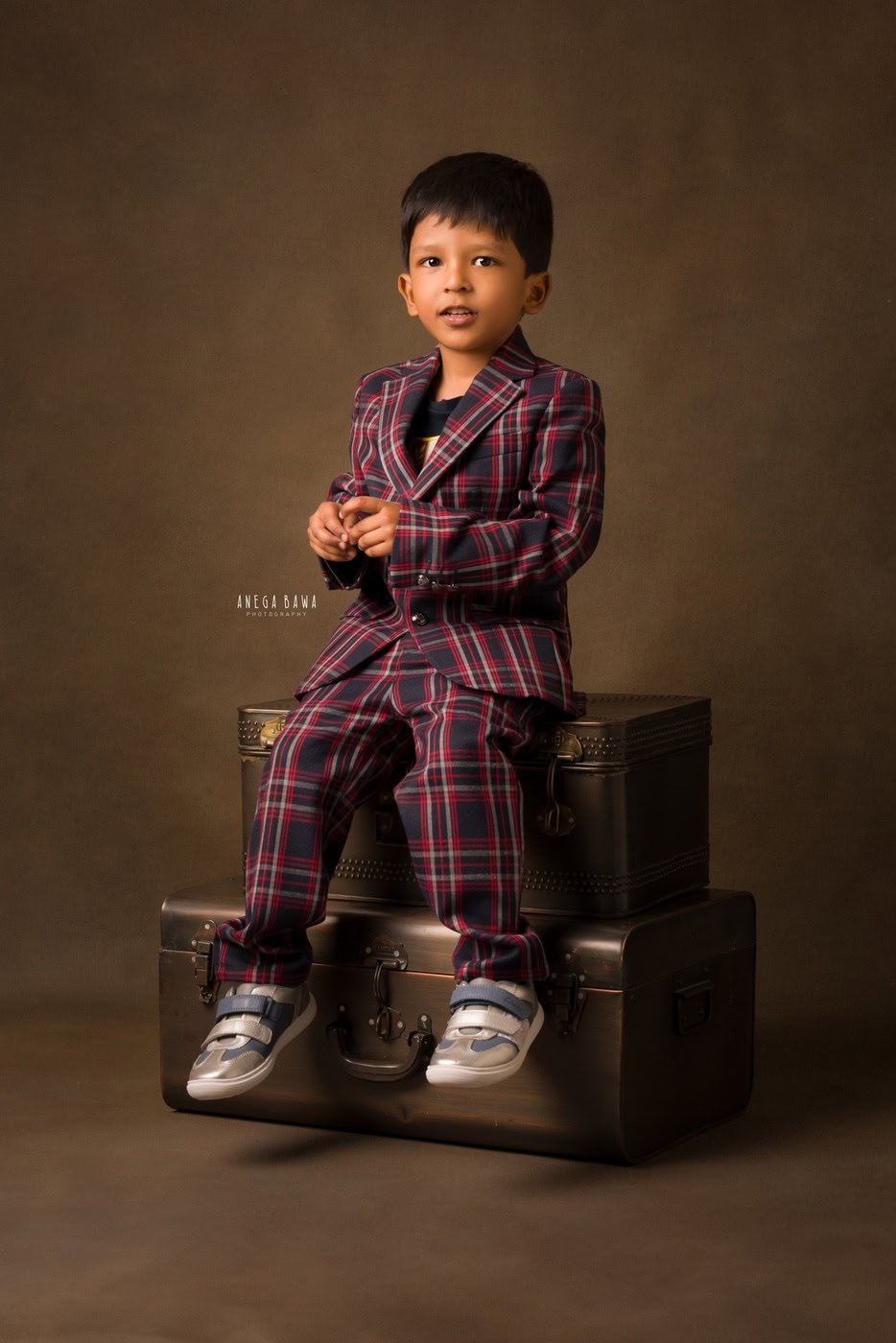 3-year-old boy posing in a check suit in a sitting pose against a brown backdrop, captured during a family photography session by Anega Bawa in Delhi, Gurgaon. His stylish outfit and confident expression showcase his adorable charm.
