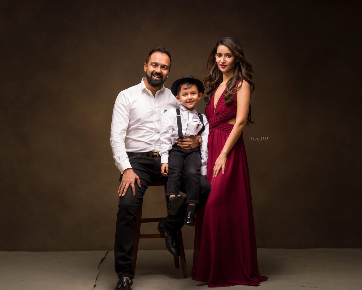 3 year boy wearing a white shirt and cute hat, posing with mom and dad against a brown backdrop, captured during a family photography session by Anega Bawa in Delhi, Gurgaon. Their joyful expressions and coordinated outfits enhance the warmth of the moment.