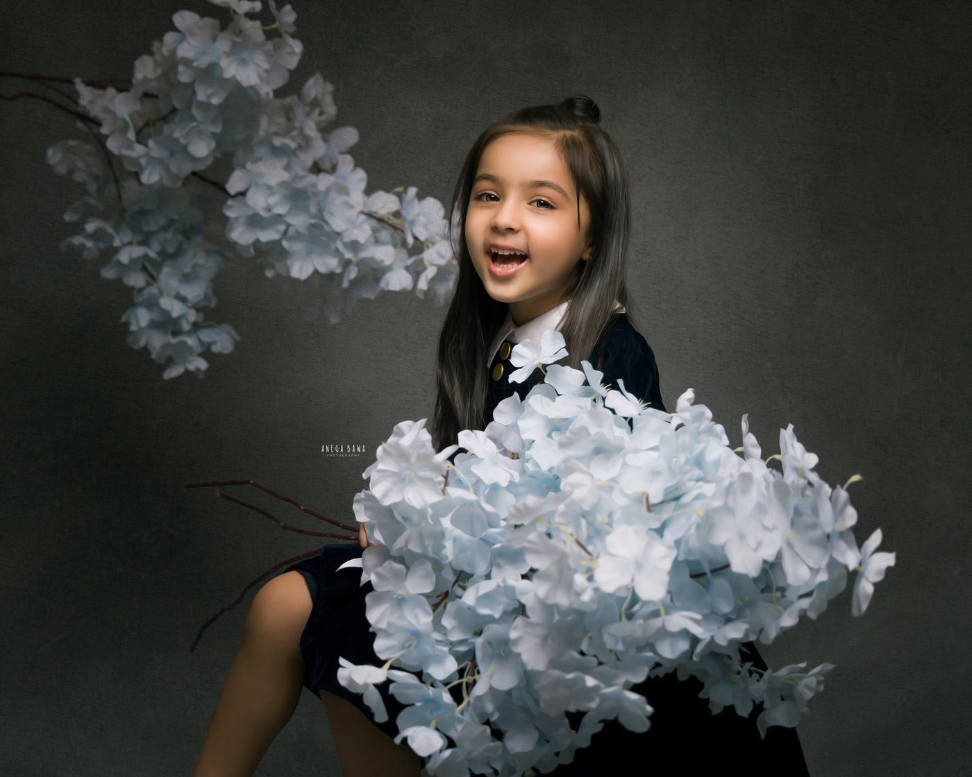 3 year girl posing cutely with paper flowers against a grey backdrop, captured during a family photography session by Anega Bawa in Delhi, Gurgaon. Her smiling pose and the vibrant flowers create a joyful and playful atmosphere.