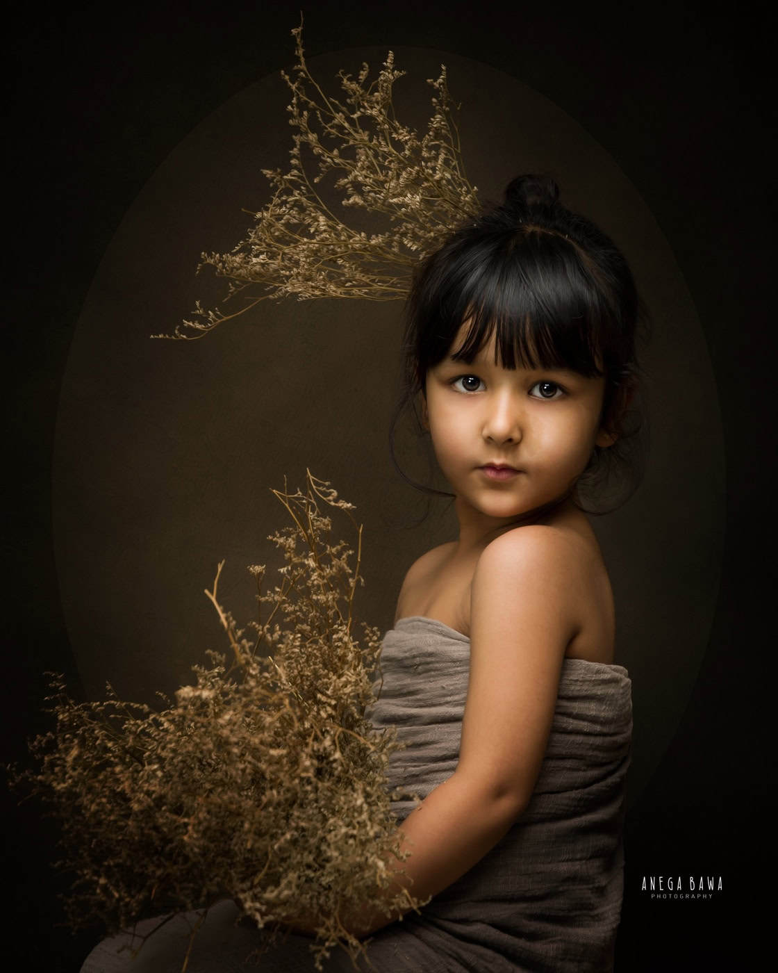 3 year girl wearing a grey wrap, posing cutely with golden shrubs against a brown backdrop, captured during a family photography session by Anega Bawa in Delhi, Gurgaon. The rich colors and playful pose create a warm and inviting atmosphere.