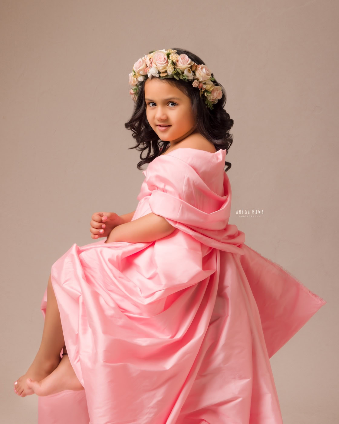 3-year-old girl wearing a pink wrap dress and a floral tiara band, posing cutely against a beige backdrop, captured during a family photography session by Anega Bawa in Delhi, Gurgaon. Her adorable pose and bright smile highlight her playful spirit.