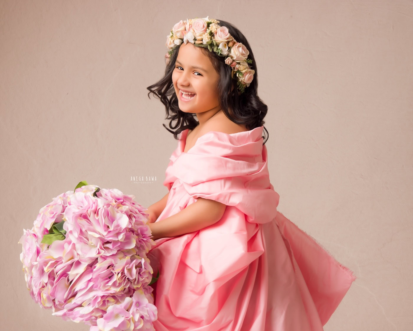 3-year-old girl wearing a pink wrap dress, holding a bunch of flowers and wearing a tiara band against a beige backdrop, captured during a family photography session by Anega Bawa in Delhi, Gurgaon. Her charming smile and delightful pose reflect her sweetness and joy.