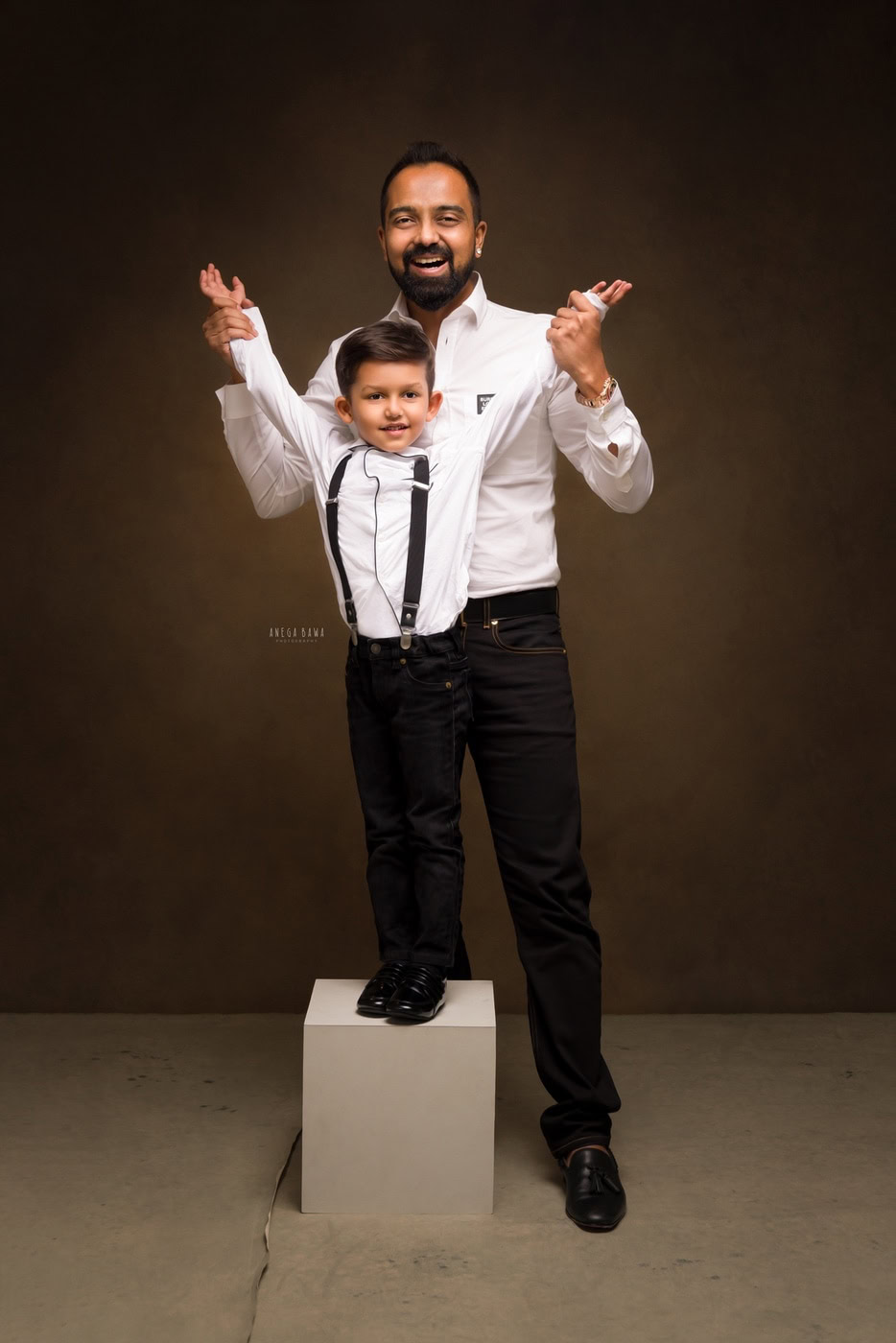 3 year boy wearing a white shirt and black trousers, posing with dad against a brown backdrop, captured during a family photography session by Anega Bawa in Delhi, Gurgaon. Their coordinated outfits and joyful expressions create a heartwarming moment.