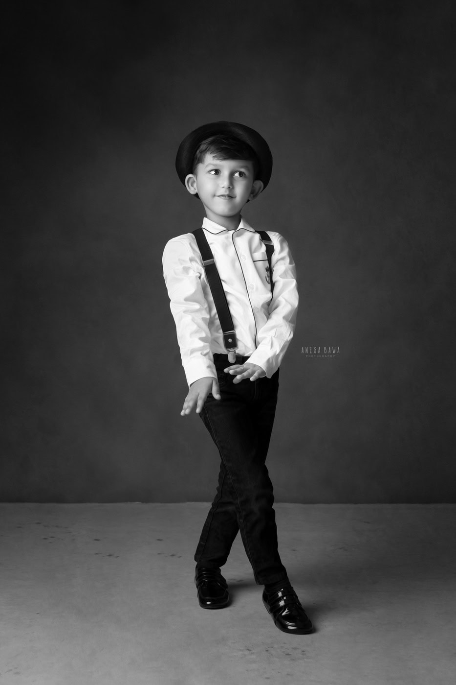 3 year boy wearing a white shirt and black trousers, posing cutely against a grey backdrop in a black-and-white family photography session by Anega Bawa in Delhi, Gurgaon. The classic tones enhance his playful charm and style.