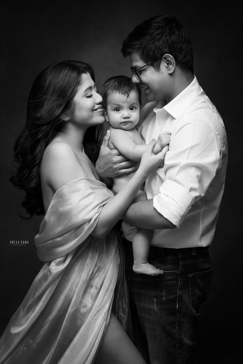 6 month boy posing cutely with mom and dad in a hugging pose against a black backdrop, captured during a family photography session by Anega Bawa in Delhi, Gurgaon. Their affectionate interaction showcases the loving family dynamic.
