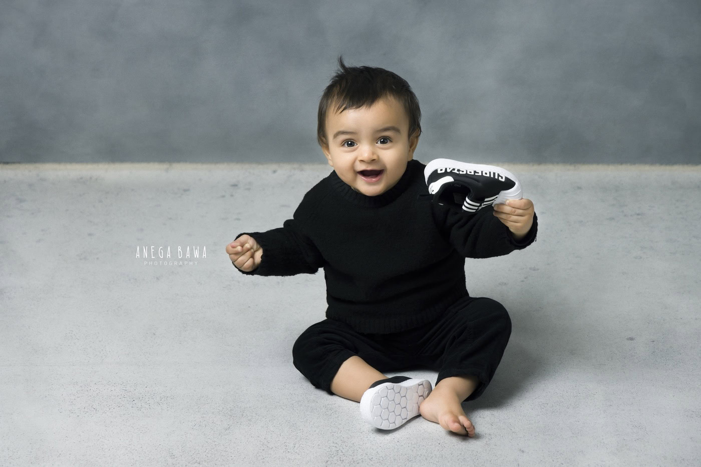Boy wearing all black, playing with his own booties against a grey backdrop, captured during a family photography session by Anega Bawa in Delhi, Gurgaon. His playful demeanor adds a charming touch to the scene.