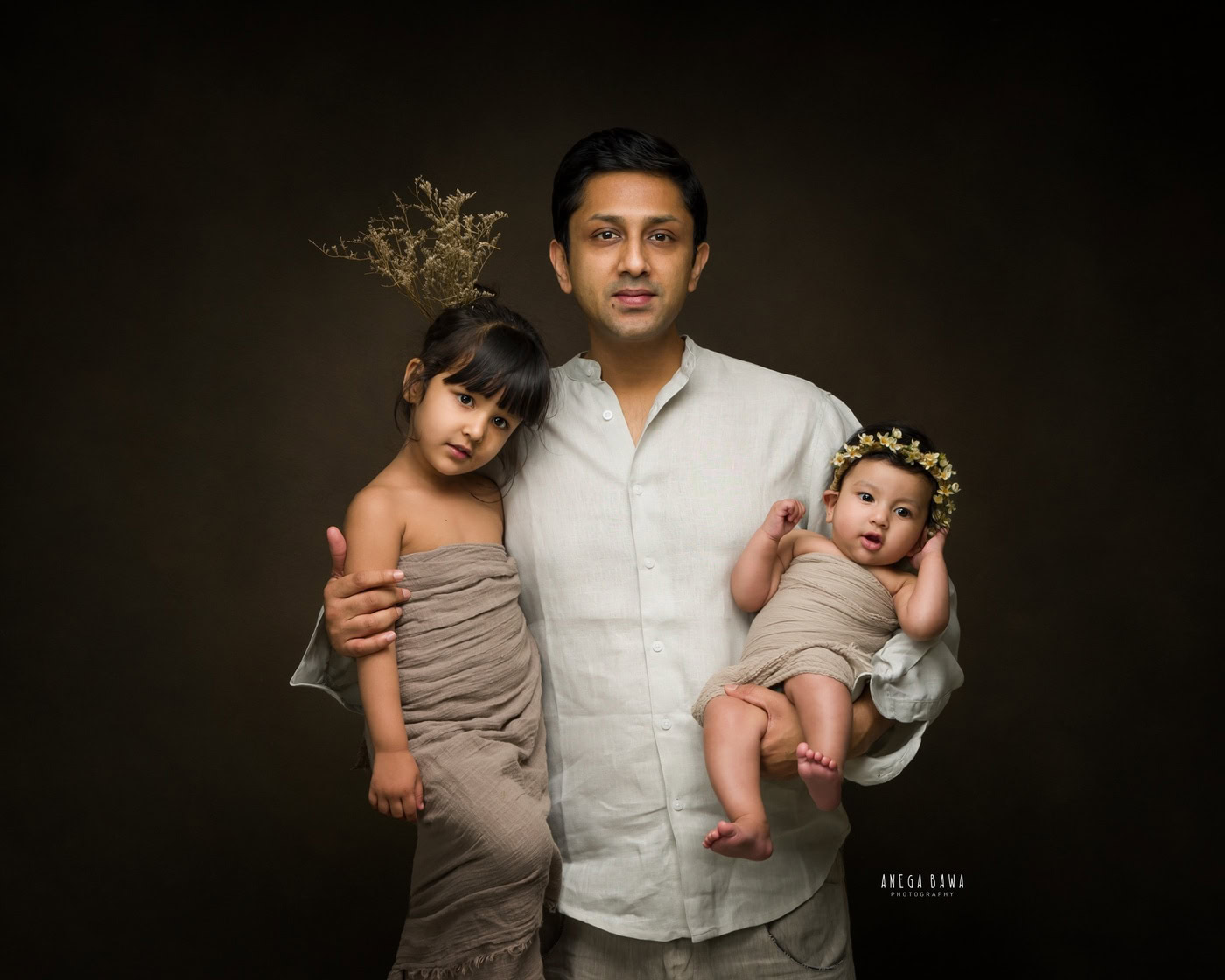 Dad holding his 1 month daughter in a beige wrap and tiara band, posing with his elder daughter also in a beige wrap against a brown backdrop, captured during a family photography session by Anega Bawa in Delhi, Gurgaon. Their coordinated outfits and loving poses highlight the warmth of their family bond.