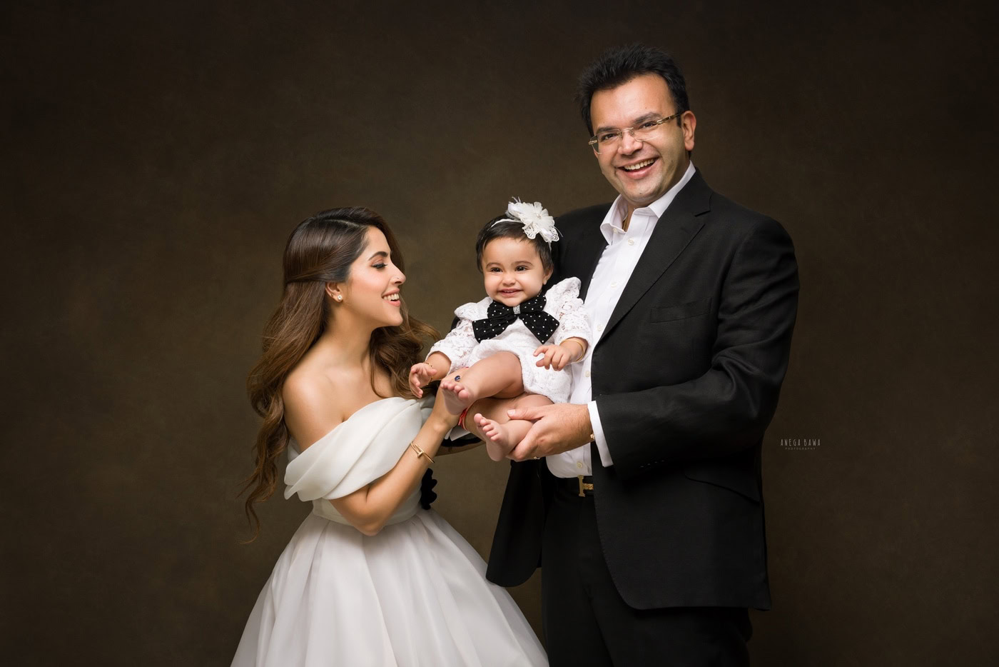 Dad holding his cute daughter in a white dress and cute headband, posing with mom against a brown backdrop, captured during a family photography session by Anega Bawa in Delhi, Gurgaon. Their joyful expressions showcase the warmth of their family connection.