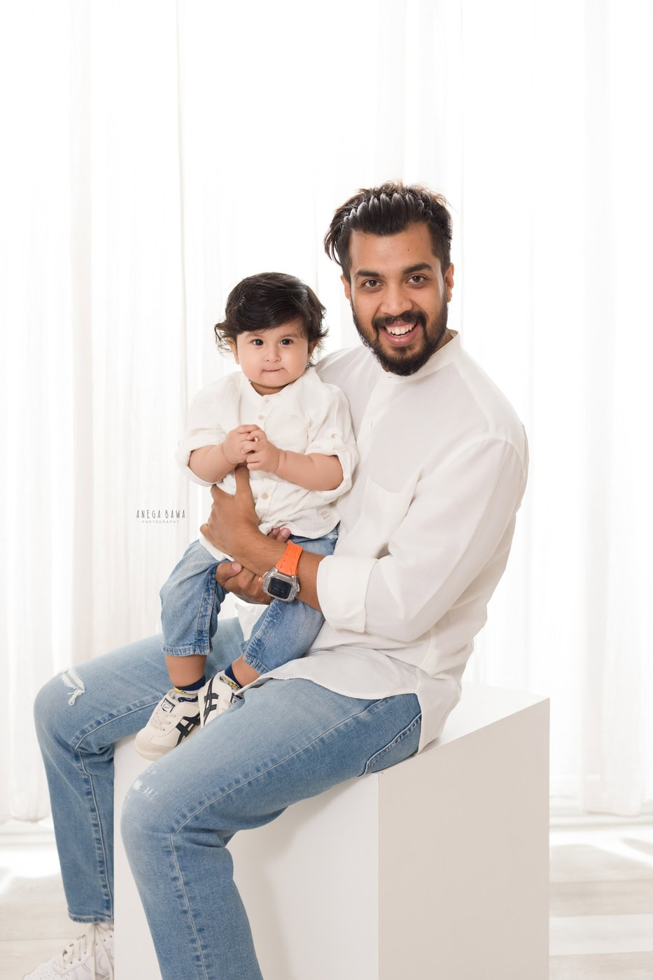 Dad holding his cute son, both twinning in white shirts and denim against a white backdrop, captured during a family photography session by Anega Bawa in Delhi, Gurgaon. Their coordinated outfits and joyful expressions highlight their special bond.