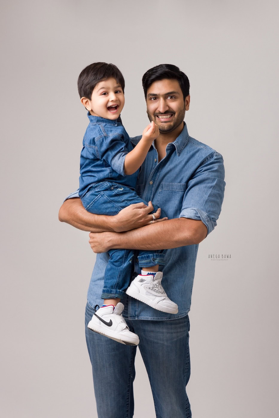 Dad holding his son, both wearing denim shirts in a smiling pose against a grey backdrop, captured during a family photography session by Anega Bawa in Delhi, Gurgaon. Their cheerful expressions emphasize the joyful connection they share.
