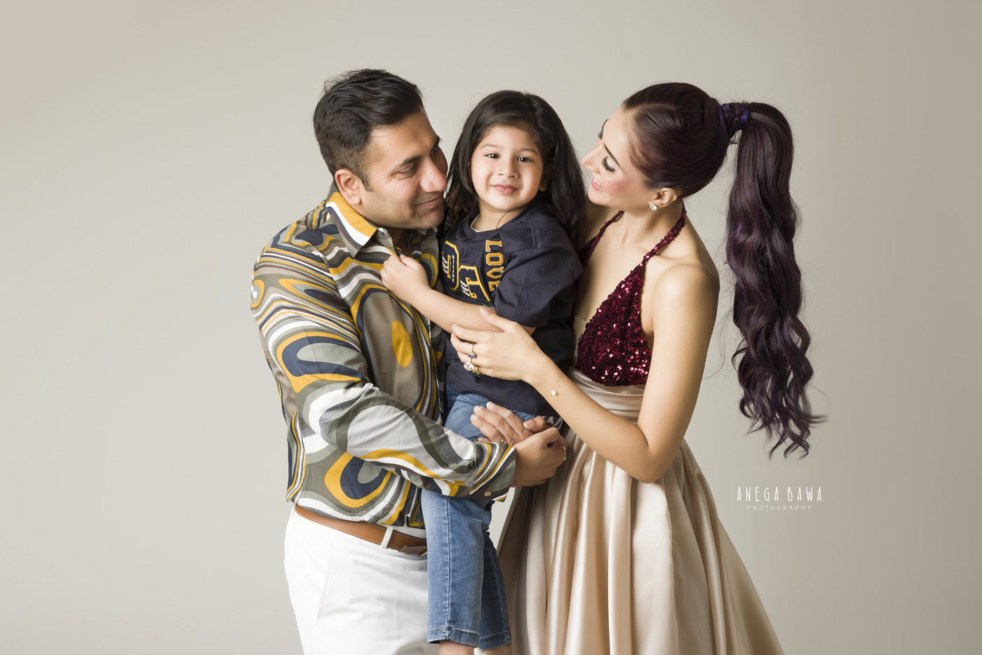 Dad holding his son while posing with mom in a smiling pose against a light beige backdrop, captured during a family photography session by Anega Bawa in Delhi, Gurgaon. Their joyful expressions and close-knit interaction beautifully showcase their family love.
