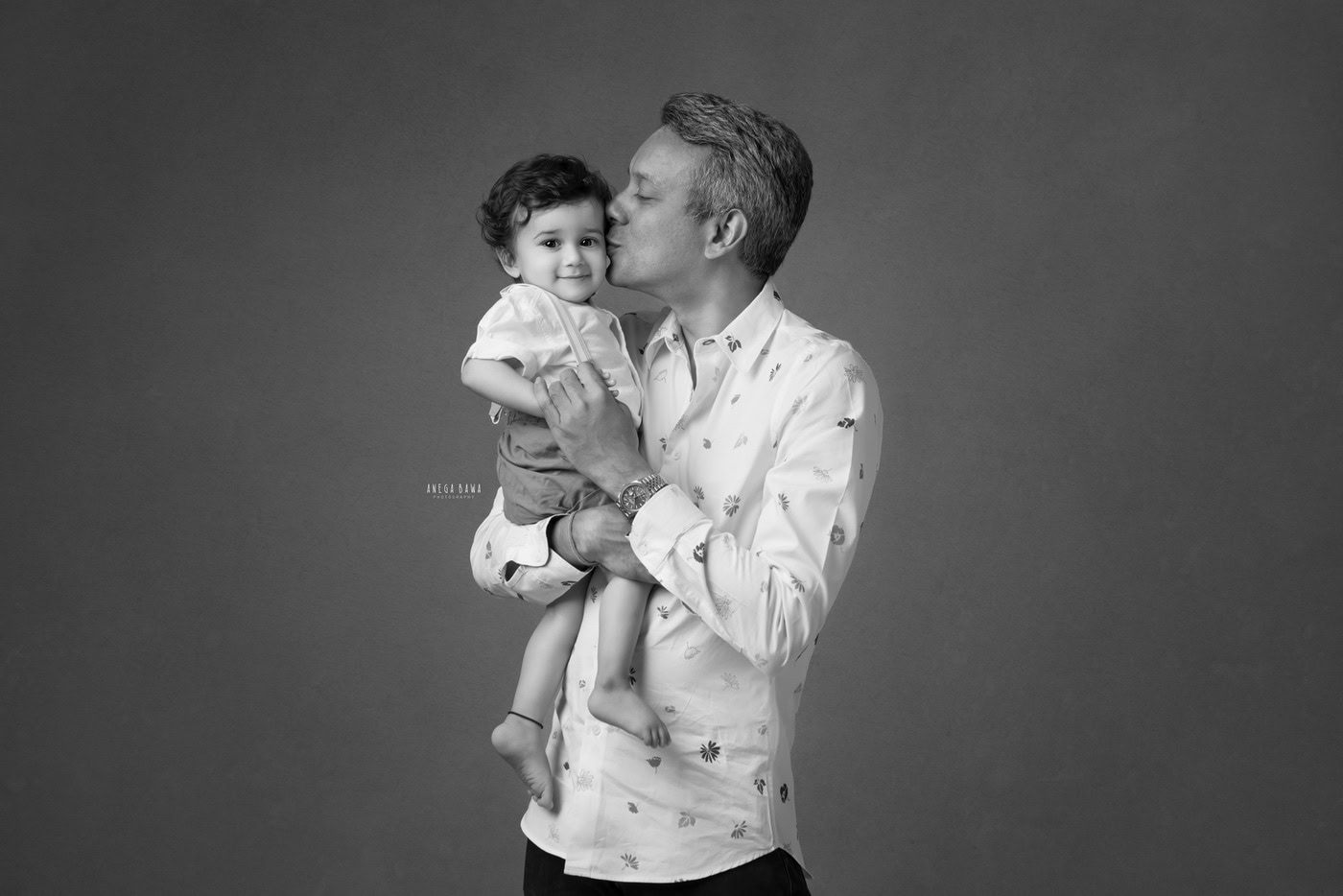 Dad kissing his cute daughter against a grey backdrop, captured during a family photography session by Anega Bawa in Delhi, Gurgaon. This tender moment beautifully showcases their affectionate bond.